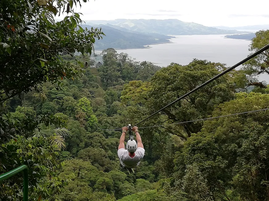 A person zip lines high above a deep green valley with a waterfall visible in the background.