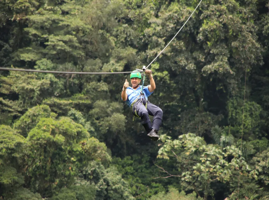 A person zip lines across a cable above a lush green tropical forest canopy.